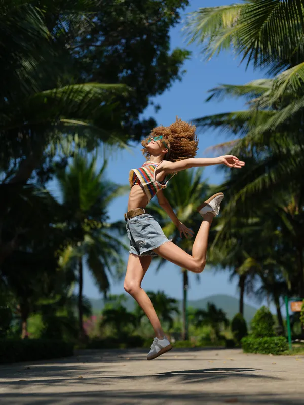 Energetic woman jumping outdoors surrounded by tropical trees, wearing casual summer clothes with joy and freedom in bright daylight.