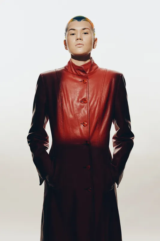 woman in long red leather coat with short hair stands confidently against white background, showcasing modern fashion and strong attitude in studio lighting.