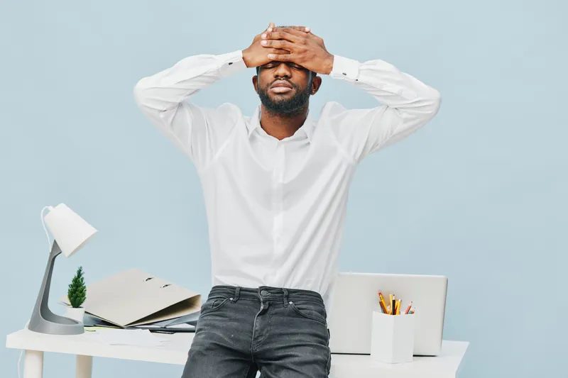 Relaxed young man with beard in white shirt sitting at office desk with laptop and stationery, covering eyes with hands, feeling tired or stressed, indoor background, professional lifestyle concept.