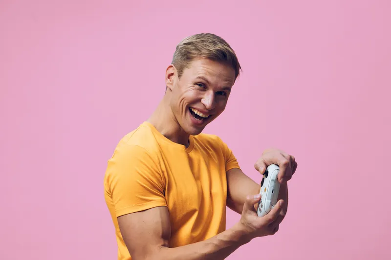 smiling young caucasian man in orange t-shirt holding gamepad enjoy digital entertainment video games isolated on pink background cheerful gamer lifestyle concept