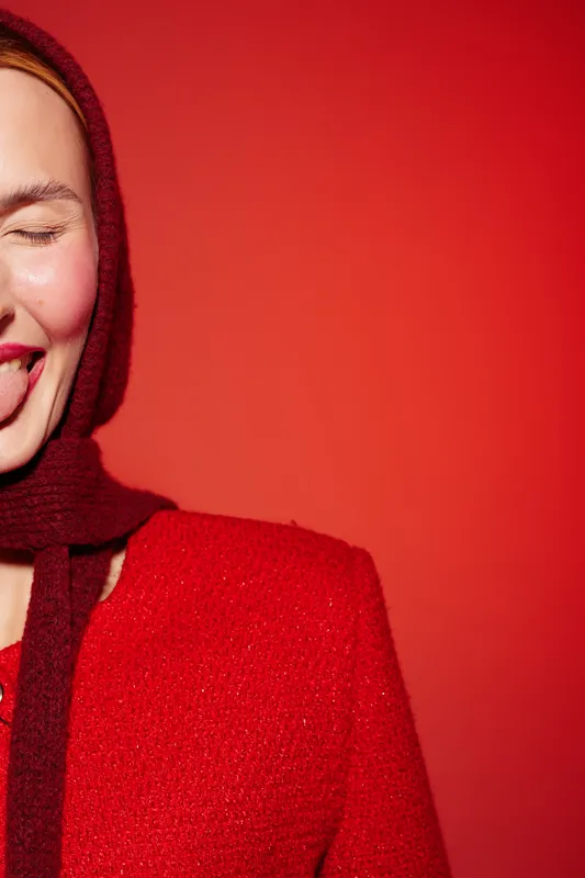 Smiling woman in red sweater and scarf with closed eyes and tongue out, posing against red background. Concept of joy, playfulness, and vibrant fashion portrait with warm tones.