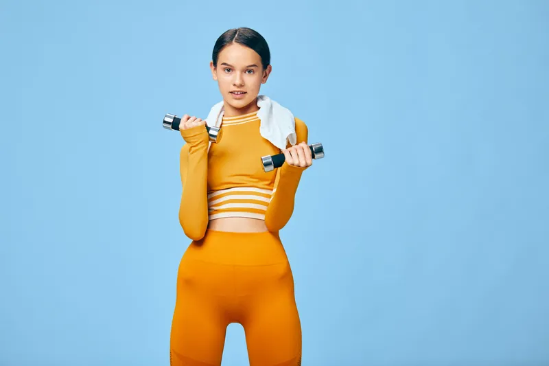 Fitness themed portrait of a woman in orange activewear lifting dumbbells with a towel over her shoulders against a blue studio backdrop, showcasing focus, strength and determination during a workout.