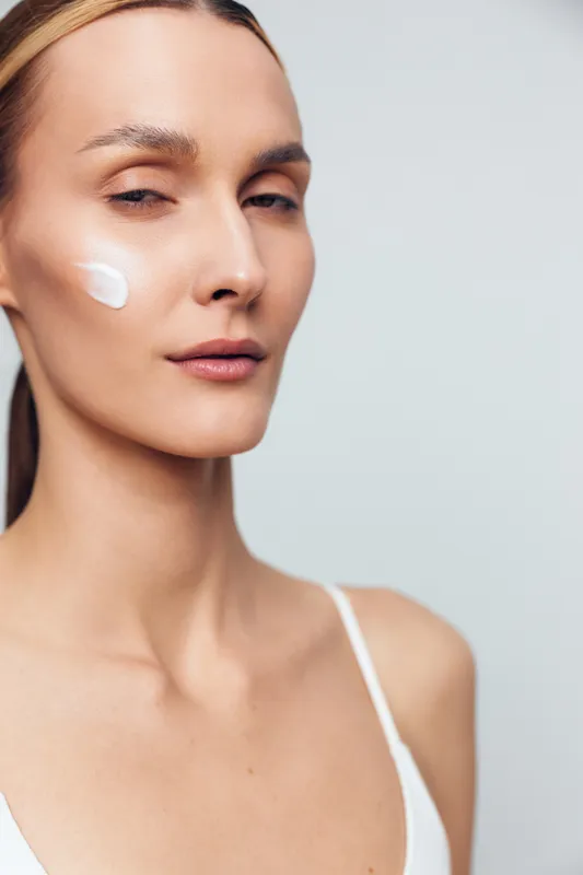 A young woman applies face cream on her cheek with a calm expression. She wears a white tank top against a neutral background. Concept for skincare, beauty, and self-care routines.