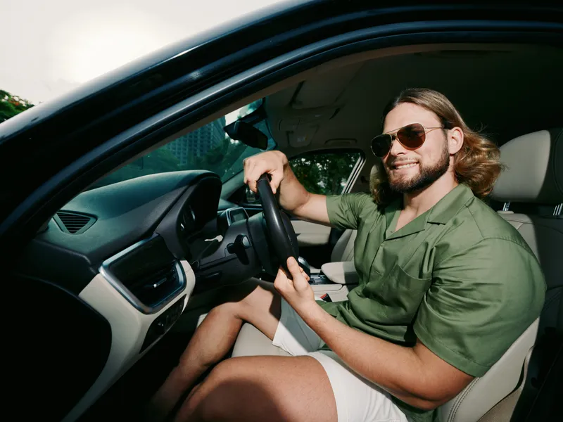 Smiling man with sunglasses driving a modern car, relaxed pose, casual green shirt, hands on steering wheel, sunlight through window, candid automotive portrait.