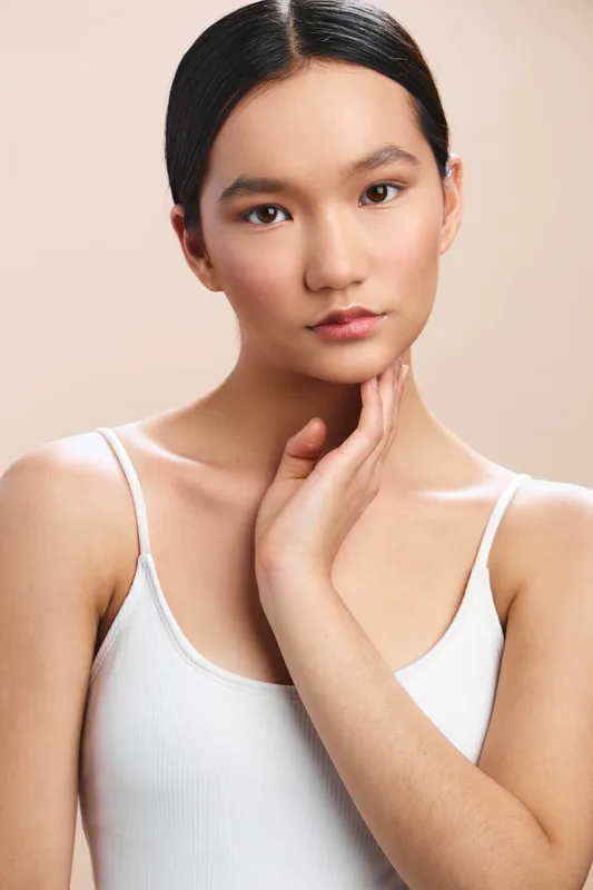 asian woman poses calmly in a simple studio setting, wearing a white tank top, soft lighting, and a serene expression that conveys poise and focus.
