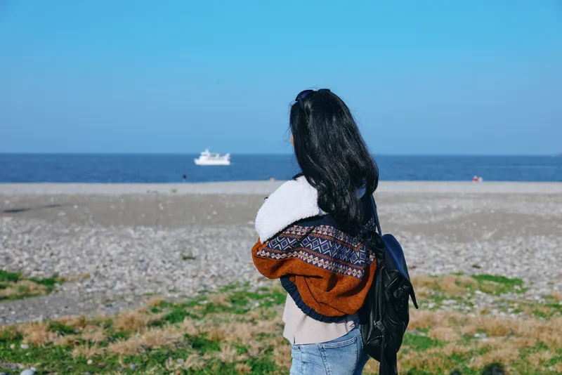 Beach scene with a woman facing the sea, wearing a scarf and denim jacket, standing on a pebble shore under a clear blue sky, conveying calm coastal atmosphere and everyday travel mood
