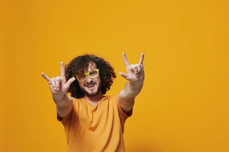 Young man with curly hair in yellow t shirt and glasses making hand gestures on vibrant yellow background, expressing joy and positivity