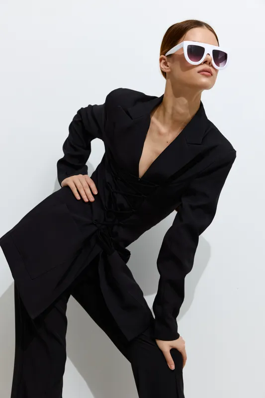 Fashion woman posing with white sunglasses, wearing black stylish outfit against minimalist light background, showcasing modern beauty and confidence.