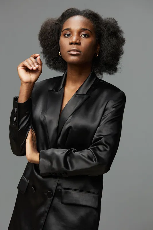 Confident Black woman in elegant satin blazer with natural hairstyle, posing against a minimalistic gray background for fashion or professional themes