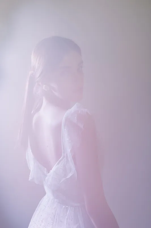 Young woman in a white dress poses with her back turned in soft light. The image highlights beauty and fashion with a dreamy, delicate atmosphere and smooth skin texture.