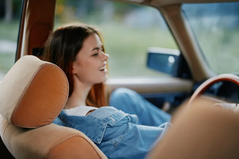 Young woman smiling in a vintage car, wearing casual denim, conveying a carefree summer spirit with a blurred green background.