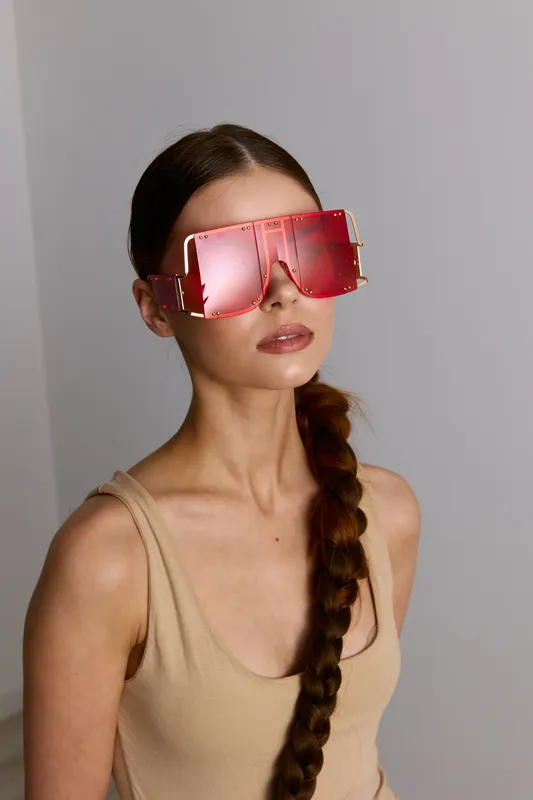woman with long braid wearing oversized pink sunglasses and beige tank top posing indoors with neutral background, modern fashion and summer style portrait