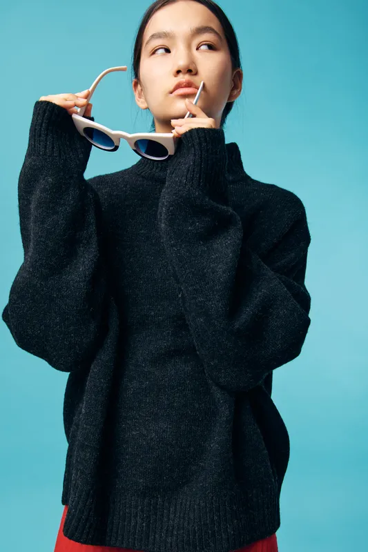 Young woman wearing black sweater posing against blue background holding white sunglasses and looking thoughtfully to the side in studio portrait with casual style.