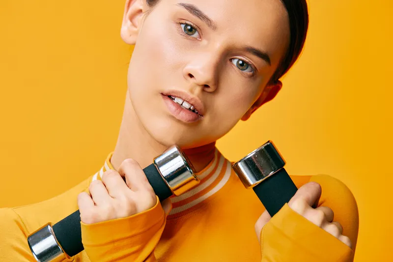 Closeup portrait of a young woman fitness, sport model in a yellow workout top holding chrome dumbbells, sweaty glowing skin and focused expression against a bold yellow studio background.
