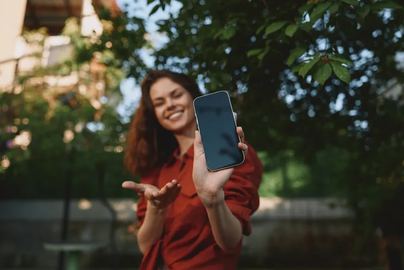 Young woman with curly hair holds a smartphone toward the camera, smiling in a sunny park as she records a casual selfie, surrounded by green trees and warm natural light