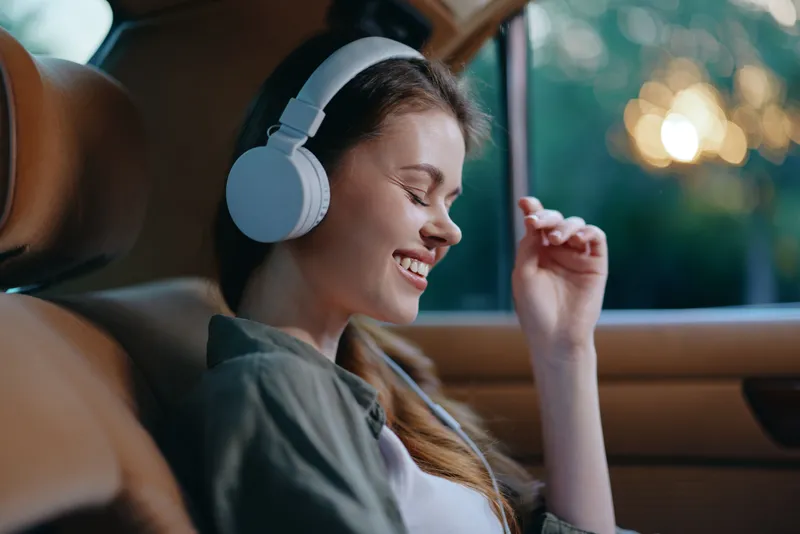Young woman enjoying music with headphones in a cozy car setting, smiling with joy as she relaxes in the sunlight, perfect for concepts of leisure, tranquility, and happiness.