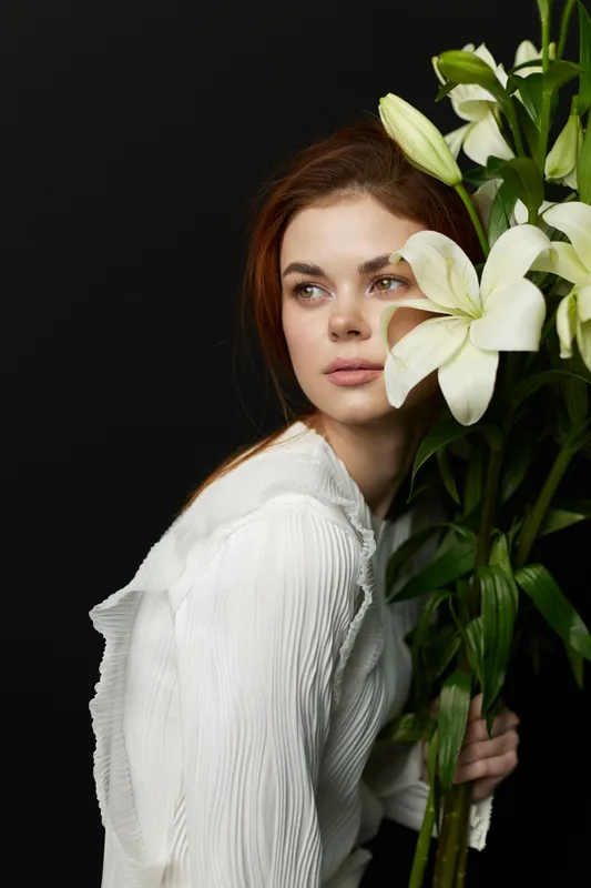 A serene woman of Eastern European descent holding white lilies, wearing a stylish white blouse, against a dark background, evoking elegance and tranquility