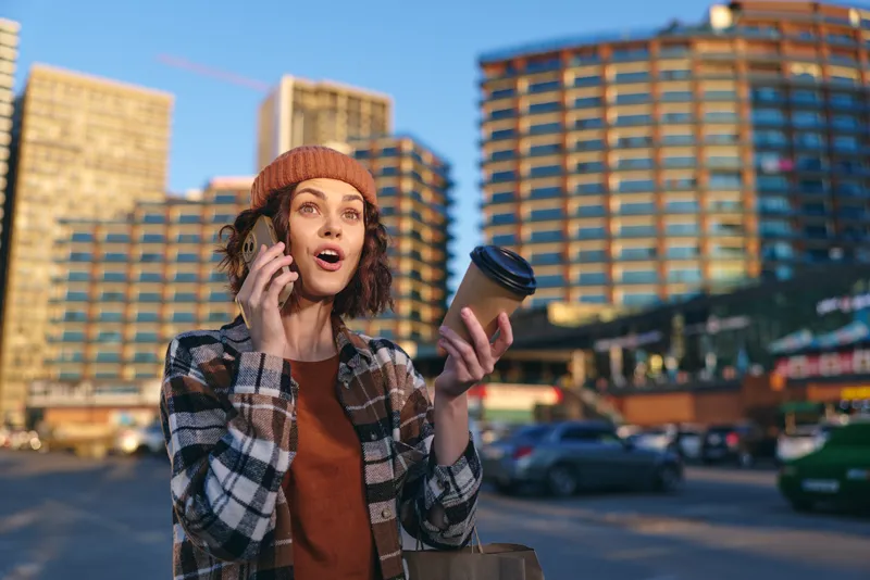 Woman holding coffee and phone on city street in beanie with surprised expression, candid urban moment. Portrait captures authenticity, candid lifestyle, golden hour glow and emotional storytelling.