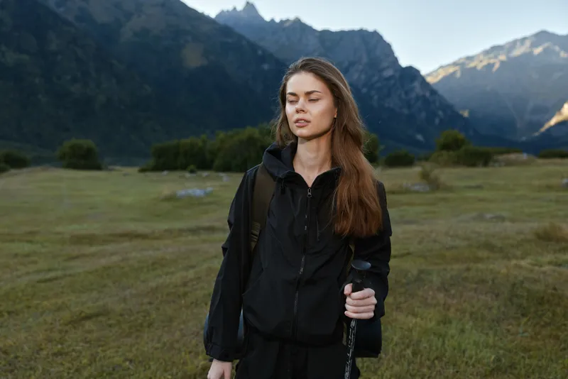 A solitary traveler walks through a grassy valley with distant mountains, wearing a dark jacket and backpack, carrying a trekking pole, and moving with calm concentration.