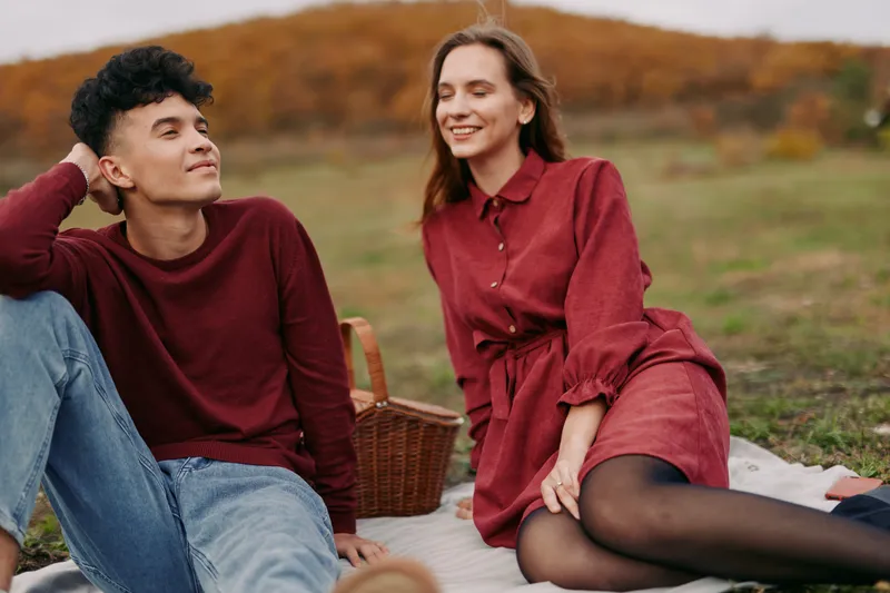 Young couple enjoying a relaxed outdoor picnic on a blanket in an autumn field, sharing genuine smiles and natural interaction that suggest authenticity and credibility in the moment.