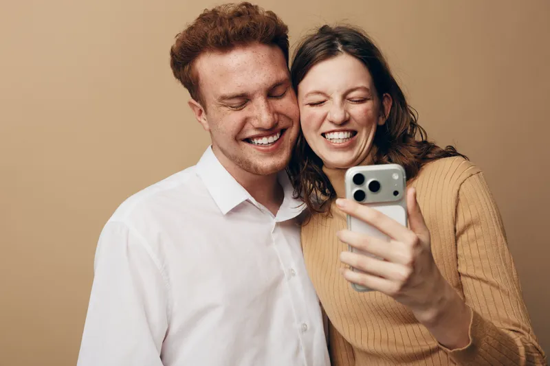 Happy young couple takes a selfie together smiling with closed eyes. Casual clothing and warm beige background create a cozy and joyful atmosphere of friendship and love.