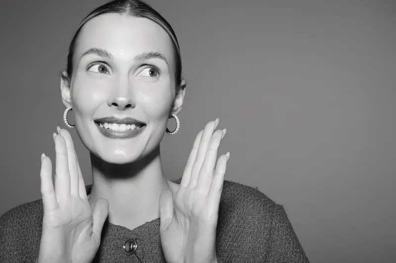 Happy white woman with light skin and slicked hair smiles and raises hands in front of grey background. Concept for joy, positivity, and natural beauty in studio portrait.