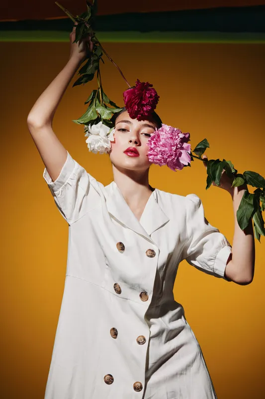 Fashion model poses with colorful flowers framing her face wearing a white dress. Bright yellow background highlights creative beauty style and floral art concept.