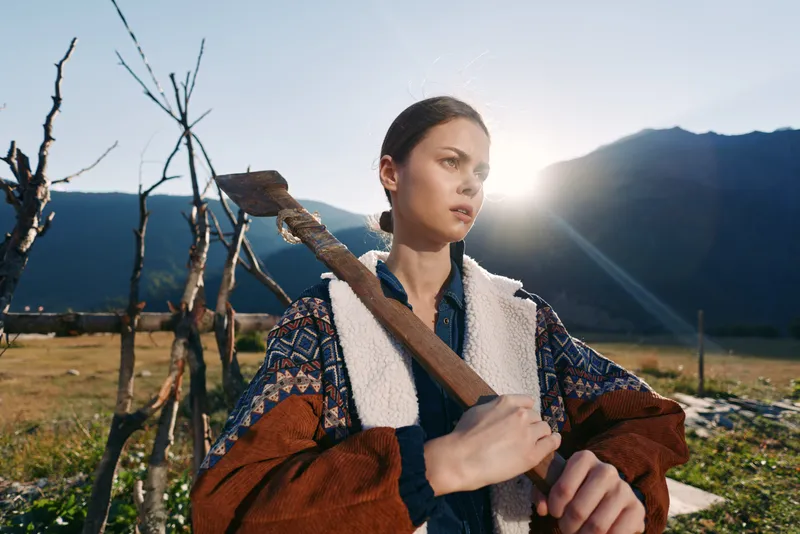 Woman with axe on shoulder on rural farm, portrait at sunrise in mountains. Outdoor worker in warm jacket, determined expression, rustic fence and field in backlit landscape.