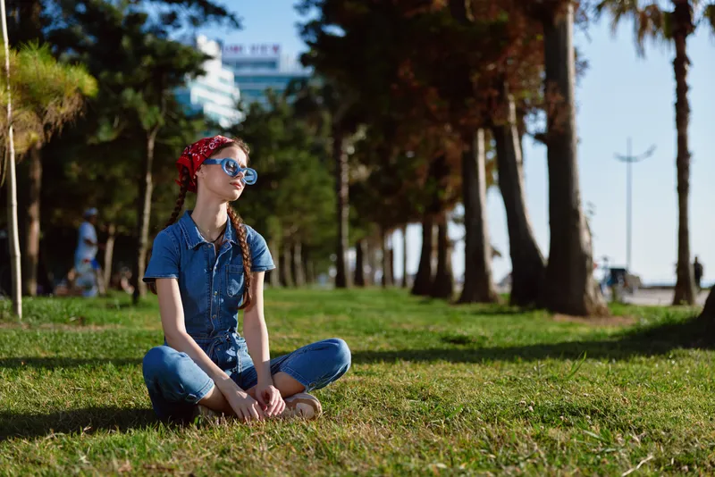 Lifestyle woman in modern boho-western denim street style sitting on grass outdoors in warm sunlight with film color tones and casual relaxed mood.
