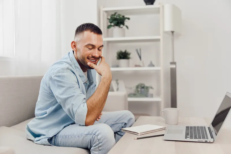 Smiling man working in a bright, cozy living room, engaging with his laptop. The scene captures a modern workspace, emphasizing productivity and comfort.