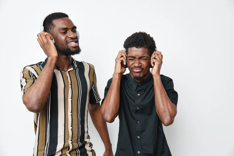 Two young black men with dark skin listening to music with headphones, smiling and enjoying sound in casual clothes on white background.