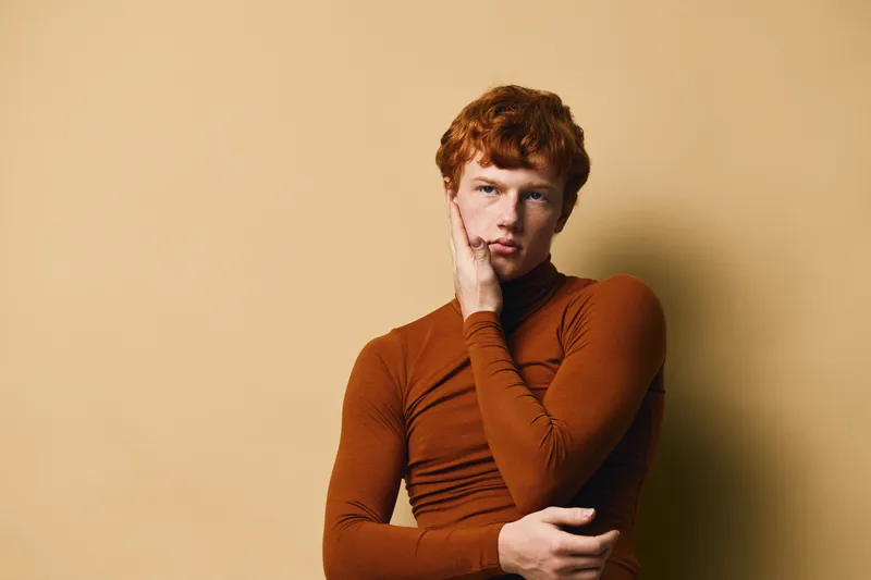 Young man with red hair and freckles wearing brown turtleneck poses with hand on face against beige background. Thoughtful expression, casual style portrait photo.