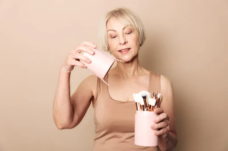 Mature woman with short blonde hair holding pink containers with makeup brushes on beige background. Concept of beauty tools, cosmetics organization, and skincare routine.