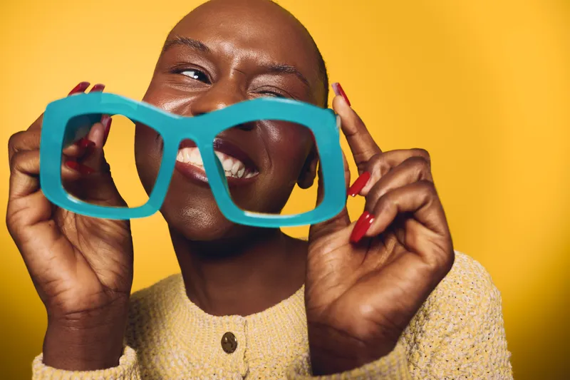 Happy dark skinned African woman with bald head holds large blue glasses frame smiling against bright yellow background. Concept of joy, fashion, and self expression with colorful style.