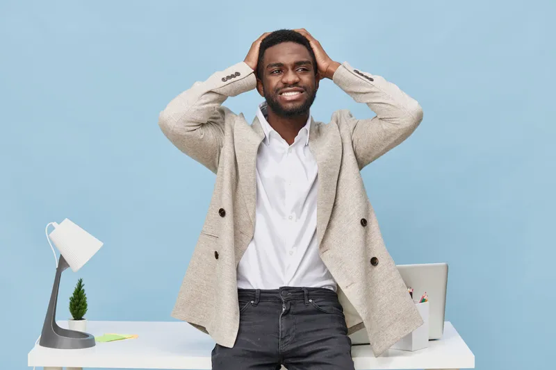 Confident smiling man with dark skin, wearing a beige blazer and white shirt, standing in front of a bright blue background, holding his head with both hands, and looking at the camera. Studio portrai