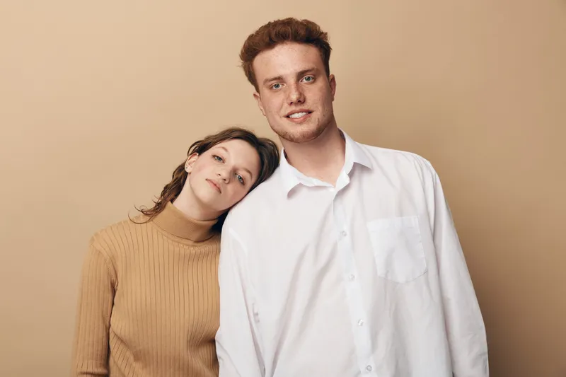 Young man and woman pose together against beige background. The woman leans her head on the man’s shoulder. Casual clothing and warm tones create a relaxed, intimate mood.