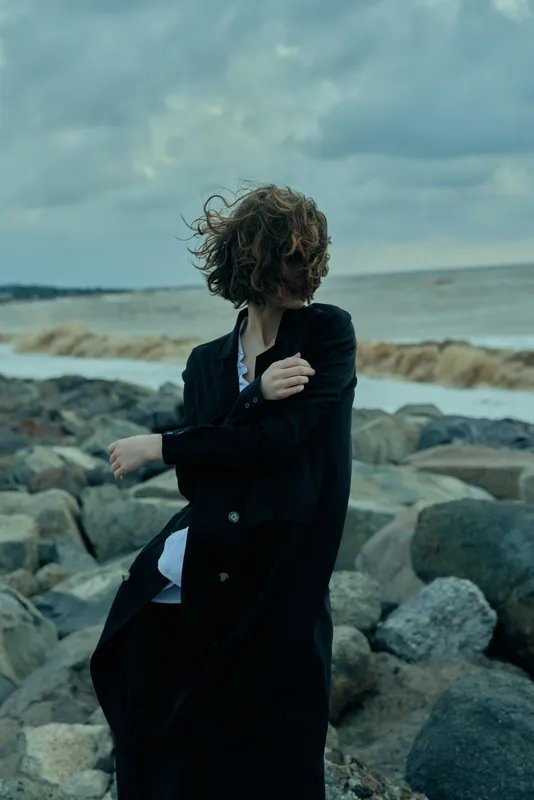 Coat wrapped against wind on a rocky shoreline with moody ocean backdrop, motion and atmosphere in clothing, solitary figure amid sea breeze and dramatic weather.