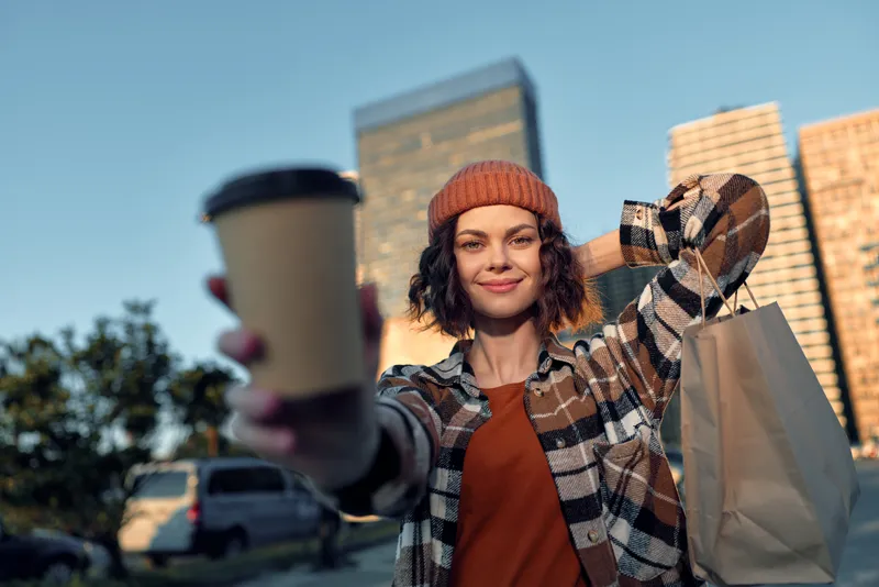 Woman holding coffee and shopping bag in urban street, smile and beanie during golden hour glow, candid lifestyle portrait with authenticity, mindful living and emotional storytelling.
