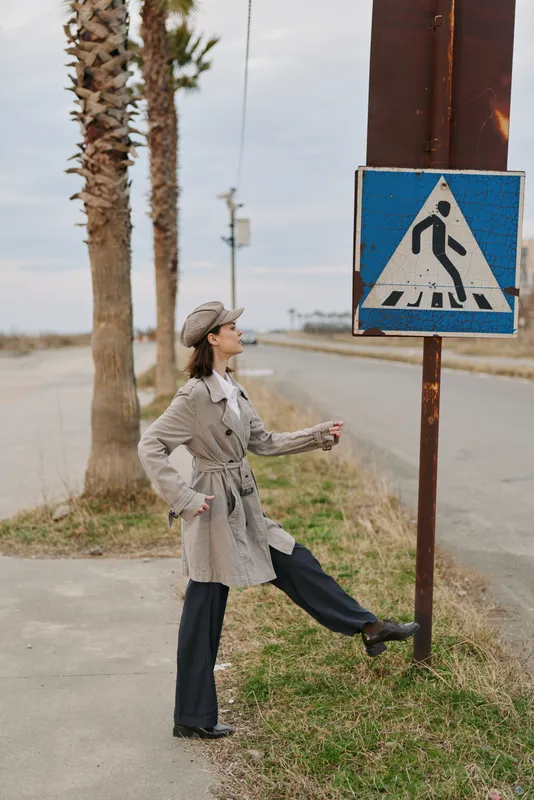 Woman in vintage outfit walks near pedestrian crossing sign on roadside with palm trees. Outdoor scene with casual fashion and urban environment under cloudy sky.