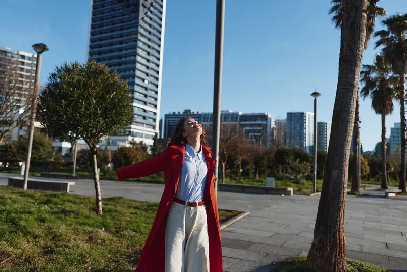 A stylish woman in a red coat stands in a sunny urban park, arms outstretched and face tilted up as tall buildings rise behind her, capturing a moment of freedom.