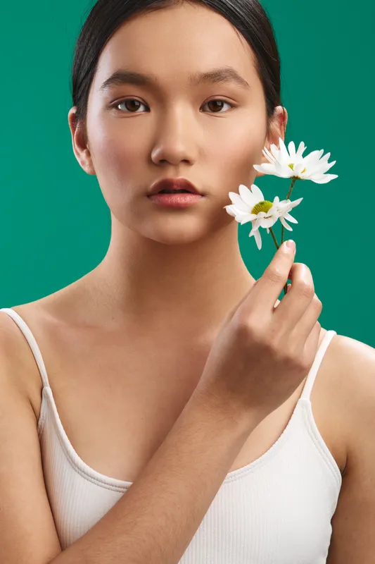 Asian woman gazes softly at the camera while holding white flowers near her face, set against a bold green backdrop to convey calm beauty and gentle natural elegance.