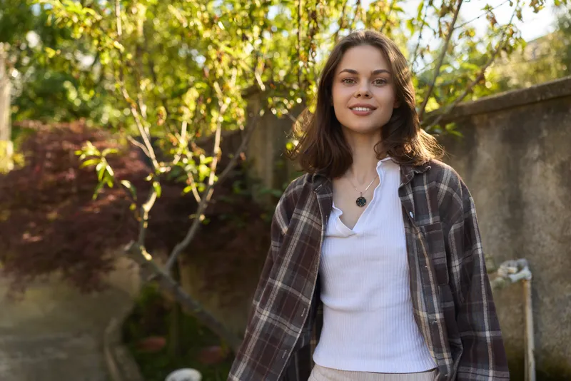 A smiling woman in a plaid jacket stands in a sunlit garden, capturing casual lifestyle in an outdoor setting. A confident portrait of style and fashion, presenting everyday elegance