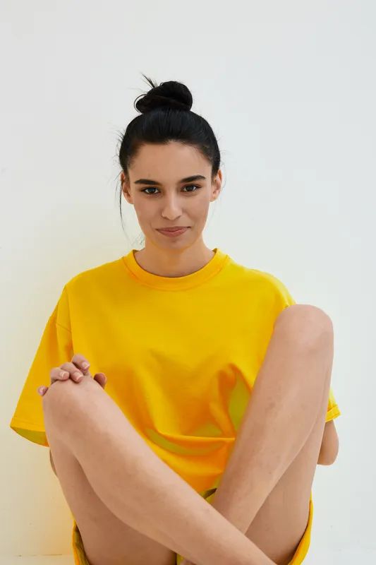 young woman sitting casually indoors wearing a bright yellow t-shirt with a neutral expression and hair tied in a bun against a plain white background