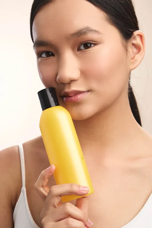 Asian woman holds a yellow cosmetic bottle in a clean studio setting, presenting a calm, confident look. The image conveys skincare focus and modern beauty routines with a soft, flattering light.