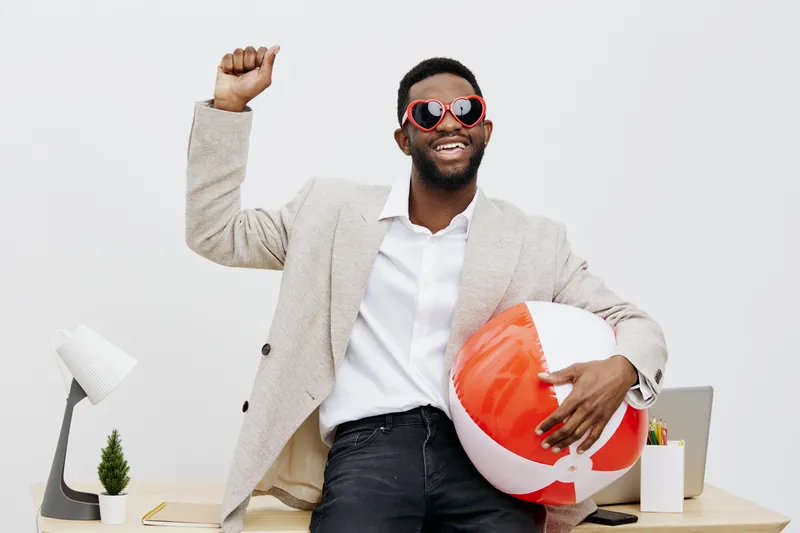 Cheerful young man with dark skin wearing beige blazer and red sunglasses holding beach ball in bright white modern office interior with minimal decor, celebrating success and happiness. People lifest