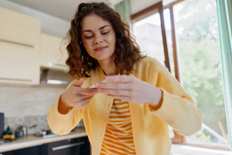 Smiling woman with curly hair looking at smartphone in a cozy home kitchen, casual everyday moment, warm natural light, modern lifestyle. She demonstrates relaxed home life in comfortable outfit.
