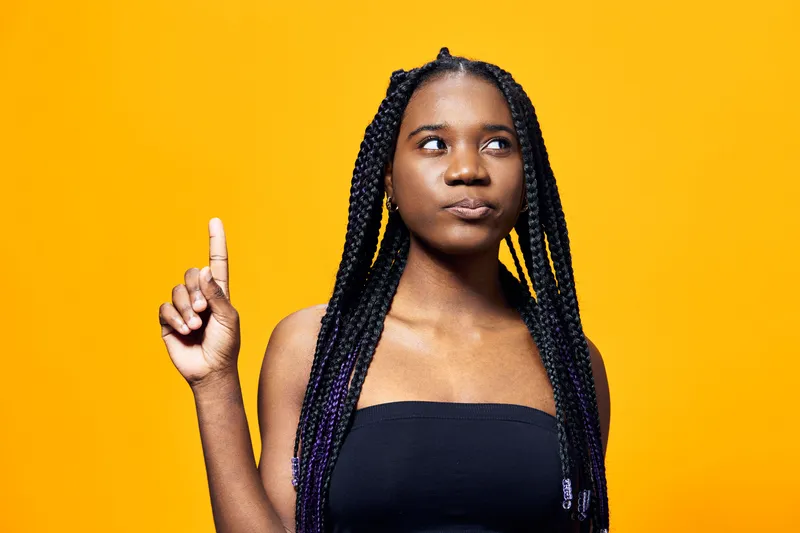 Young African woman with braided hair showing an idea gesture against a vibrant yellow background, expressing confidence and creativity