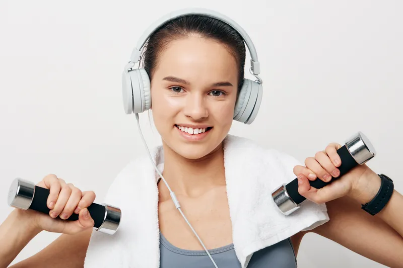 Fitness woman lifting dumbbells with a towel draped over shoulders, wearing headphones and a bright gym, capturing strength training, focus and motivation.