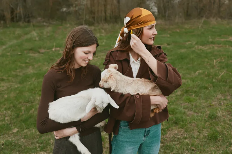 Two smiling girls in brown sweaters hold playful goat kids in a sunny field, capturing a warm lifestyle moment of kindness, nature, and play, with soft light and cozy outdoor vibes.