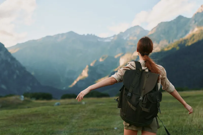 A woman hiker with a backpack stands in a meadow with arms outstretched toward distant mountains, capturing outdoor adventure, nature, and exploration in warm sunlight.